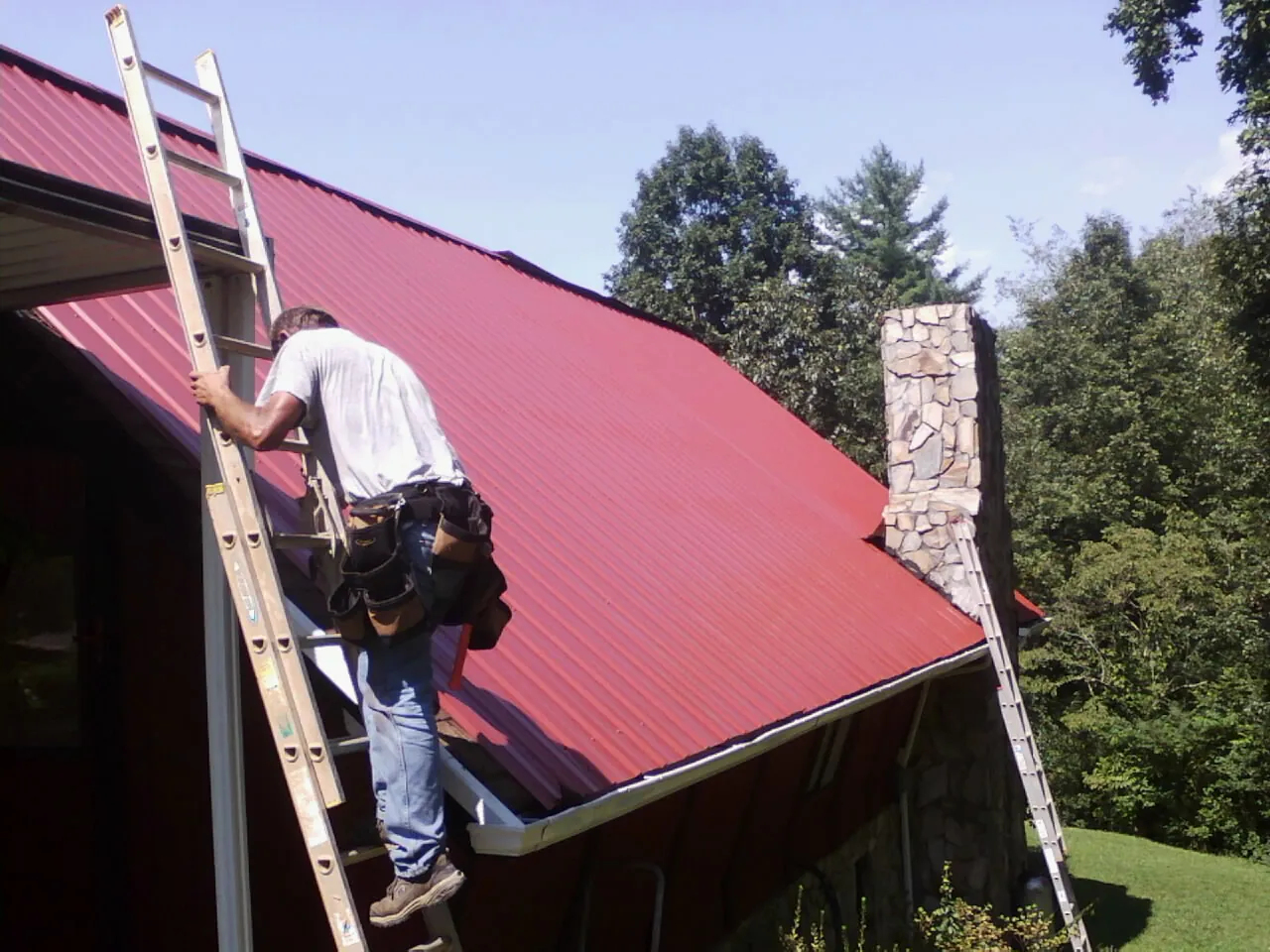Roofer on a ladder beside a red metal roof and stone chimney — Soffit & Fascia Repair by Roofing Brothers in Ludlow Falls